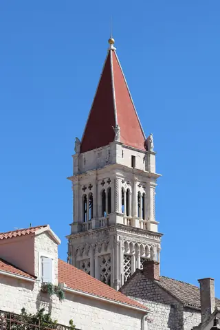 Ornate stone cathedral tower and sculpted figures above tiled roofs against blue sky.