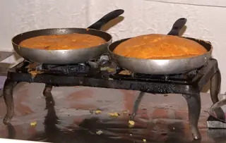 Two round golden frico preparations cook in plain metal frying pans on a working gas stove.