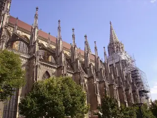 Pointed stone buttresses, windows and a scaffolded tower above leafy trees.