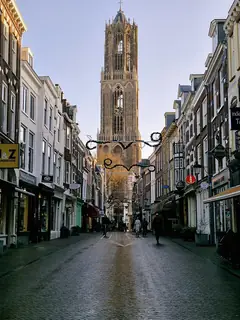 Tall Gothic stone tower above a narrow brick-paved shopping street with hanging signs, winter decorations and contextual walkers.