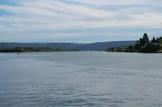 A broad river stretches toward wooded banks and low hills beneath layered clouds, with small boats and distant cranes.