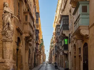 A narrow stone street with projecting enclosed balconies and a carved Saint Roque statue at the left corner.