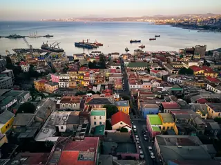 Dense hillside buildings descend toward port cranes, ships and a broad blue bay beneath a hazy sky.