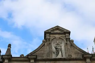 A saint holding a book beneath a stone niche and pediment, with broad blue and white cloudy sky.