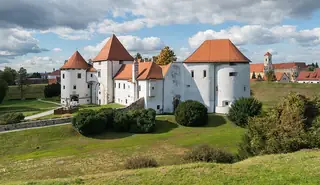 White castle walls and round towers with red roofs rise above autumn shrubs and a grassy bank under clouds.