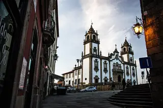 Twin church towers and an illuminated pale facade rise above a shaded Viseu street with parked cars and small pedestrians.