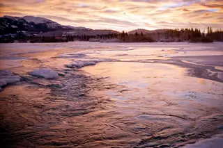 Snow and ice border open river water beneath a wooded ridge and a saturated orange-pink sky.