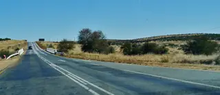 A two-lane road and low bridge cross dry grassy hills beneath a blue sky.