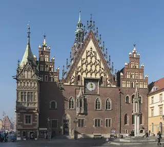 An ornate brick town hall with a clock, traceried gables and pointed turrets rises above a square with small visitors and a stone pillory.