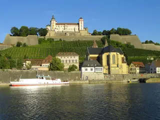 The fortress rises above a green vineyard slope, with St Burkard church, riverside buildings and a white working boat below.